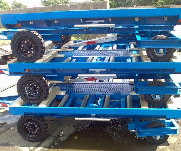 Stalowe, niebieskie wózki transportowe z kołami, ustawione jeden na drugim, z elementami kratownicy i rolkami, na zewnątrz.