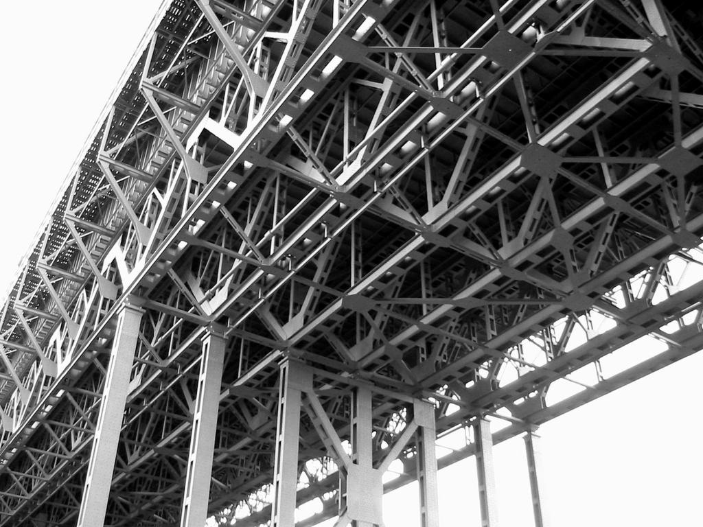 Monochromatyczne ujęcie od dołu na skomplikowaną, stalową konstrukcję hali z licznymi wzmocnieniami i kolumnami nośnymi, oświetloną z góry.