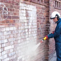Pracownik w kombinezonie ochronnym czyści czerwoną ceglaną ścianę z graffiti za pomocą myjki ciśnieniowej. Widoczny efekt czyszczenia i fragment chodnika.