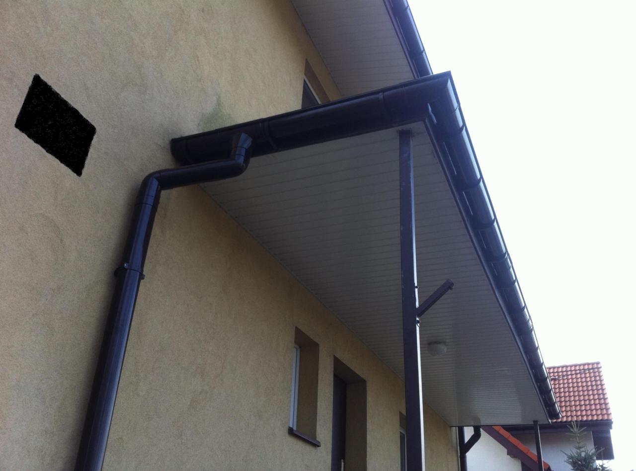 Elewacja budynku z zamontowanym systemem rynnowym w kolorze ciemnego brązu. Widoczne okna i fragment dachu sąsiedniego budynku.