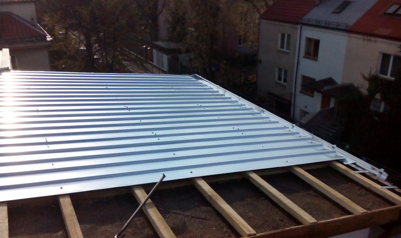 Montaż blachy trapezowej na dachu z drewnianą konstrukcją, widok z góry. W tle budynki mieszkalne i drzewa. Częściowo widoczny element mocujący.