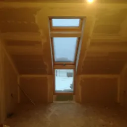 Poddasze w trakcie wykańczania: ściany i sufit pokryte płytami, widoczne łączenia szpachlowane, okno dachowe z widokiem na zaśnieżone otoczenie, surowa podłoga.