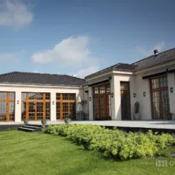 Elewacja domu z dużymi, drewnianymi oknami i drzwiami tarasowymi. Widok na zadbany trawnik i roślinność. Błękitne niebo z chmurami w tle. Charakterystyczne oświetlenie zewnętrzne.