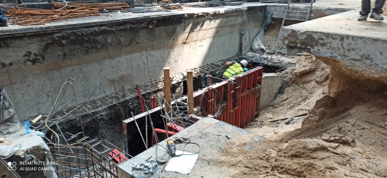 Prace ziemne przy budowie fundamentów. Widoczny pracownik w kasku i kamizelce odblaskowej montujący szalunek. W tle stalowe pręty zbrojeniowe i betonowe ściany wykopu.