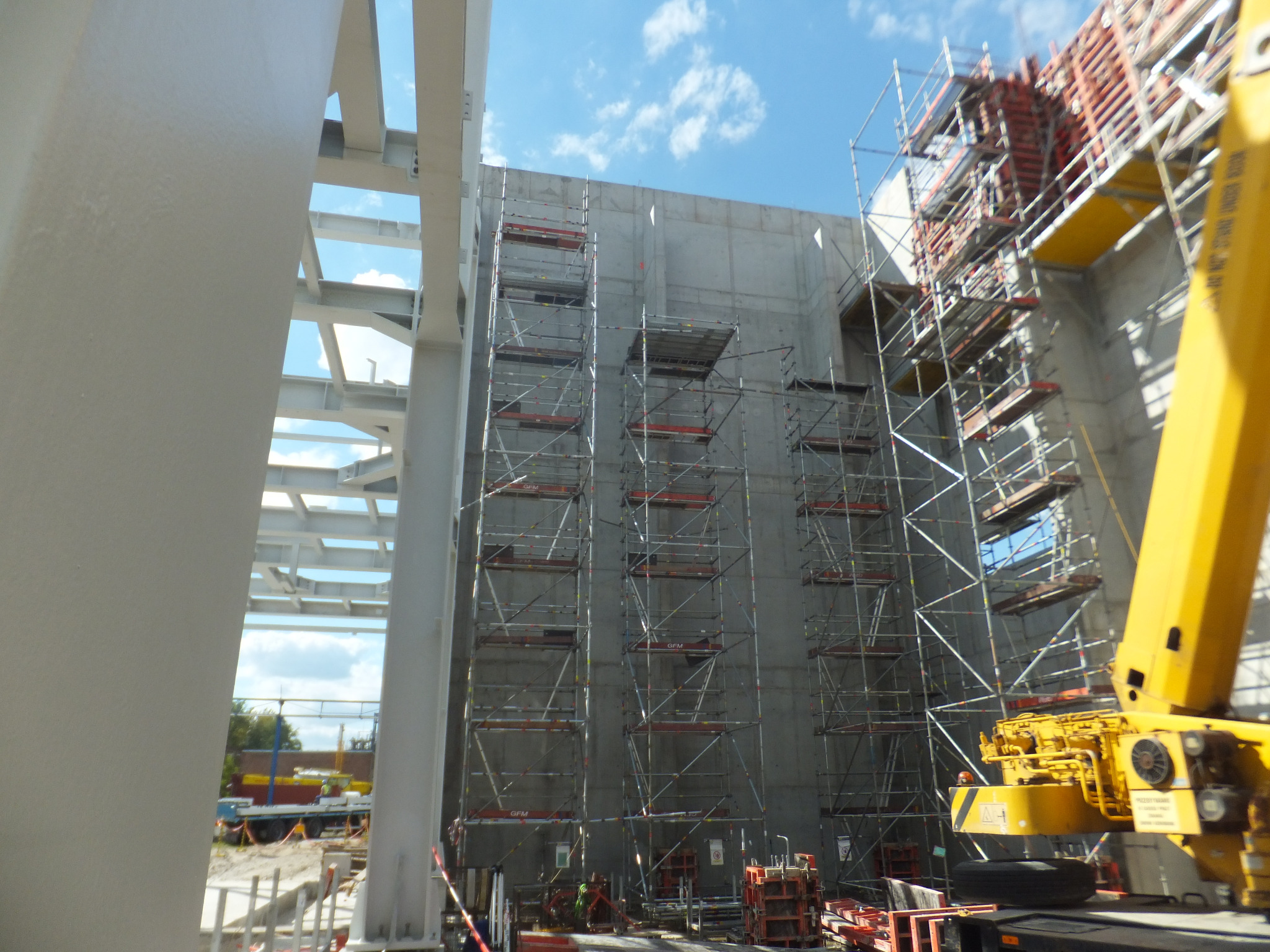 Świeżo wylany betonowy mur z rusztowaniami i żurawiem w słoneczny dzień. Widoczne szalunki i stalowe konstrukcje. Budowa budynku żelbetowego.