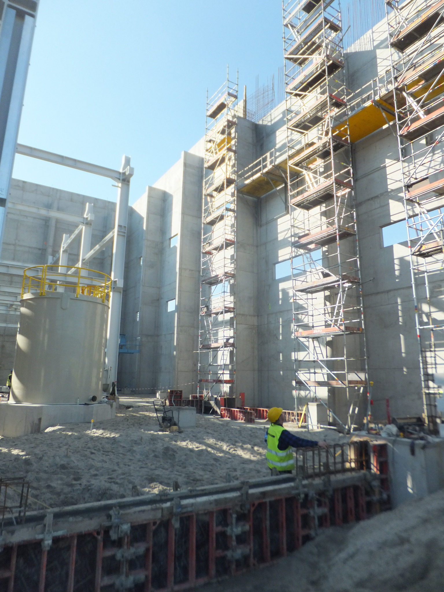 Budowa żelbetowej konstrukcji z widocznymi szalunkami i rusztowaniami. Pracownik w żółtym kasku nadzoruje prace. Jasne, naturalne światło.