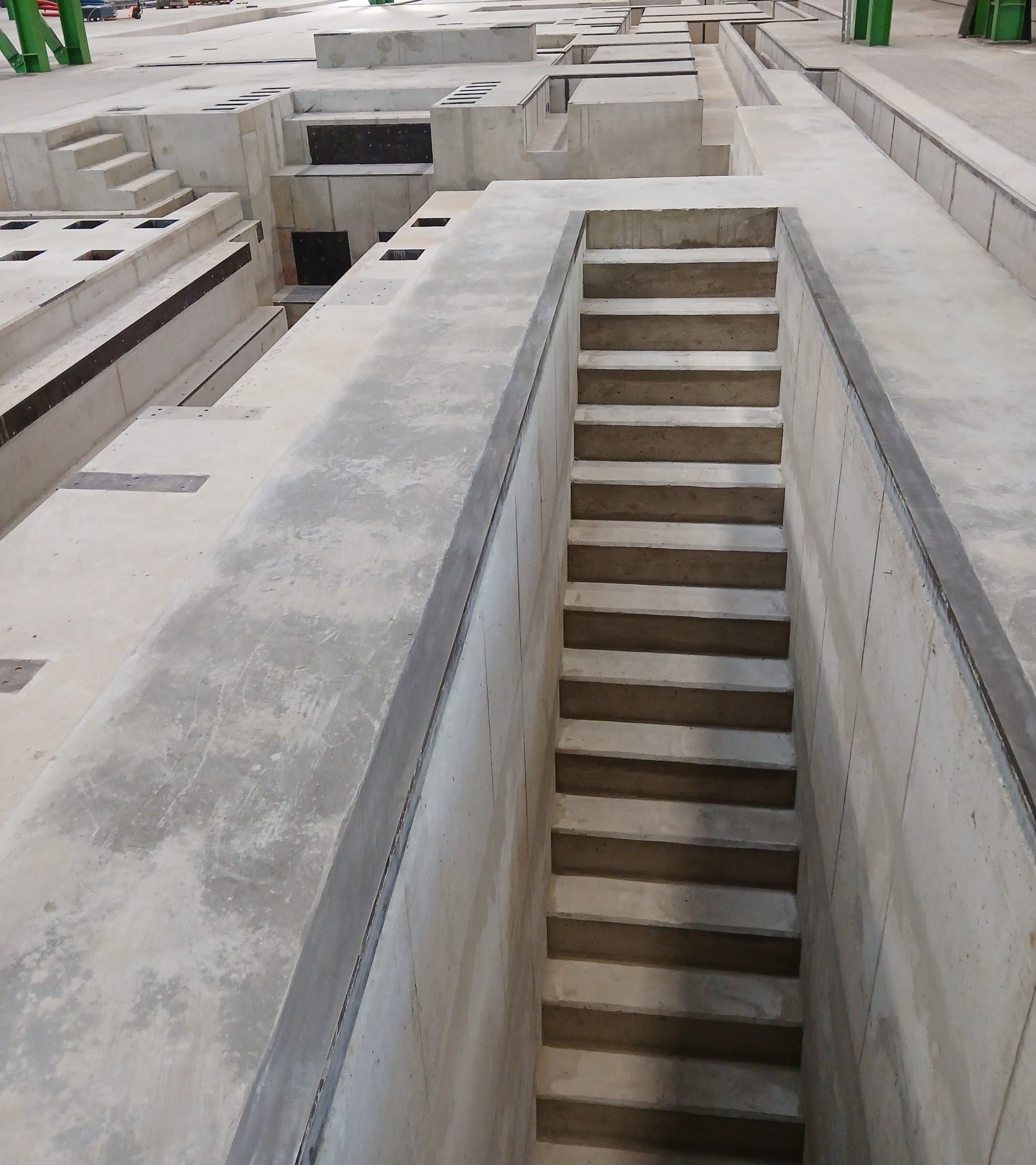 Surowe, betonowe konstrukcje w hali produkcyjnej: schody zagłębione w podłożu, elementy prefabrykowane. Widok z góry, szara kolorystyka, surowy industrialny klimat.