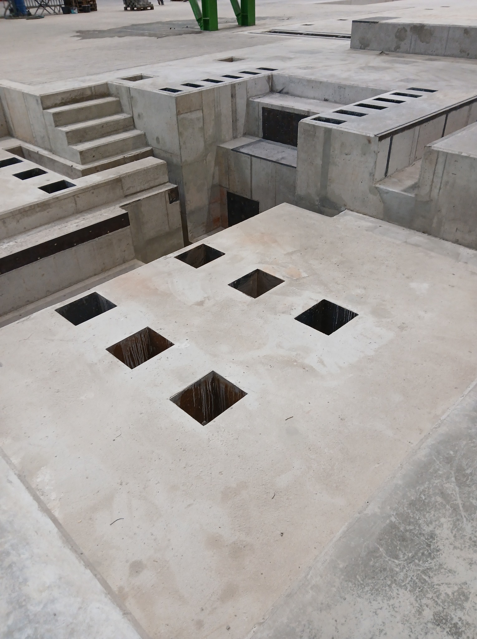 Betonowa konstrukcja z kwadratowymi otworami i schodami, surowy stan, widok z góry. Elementy konstrukcyjne w szarej tonacji, w tle fragment konstrukcji stalowej.