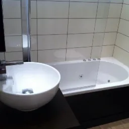 Nowoczesna łazienka z owalną umywalką na ciemnym blacie i białą wanną z hydromasażem, wykończona jasnymi płytkami.