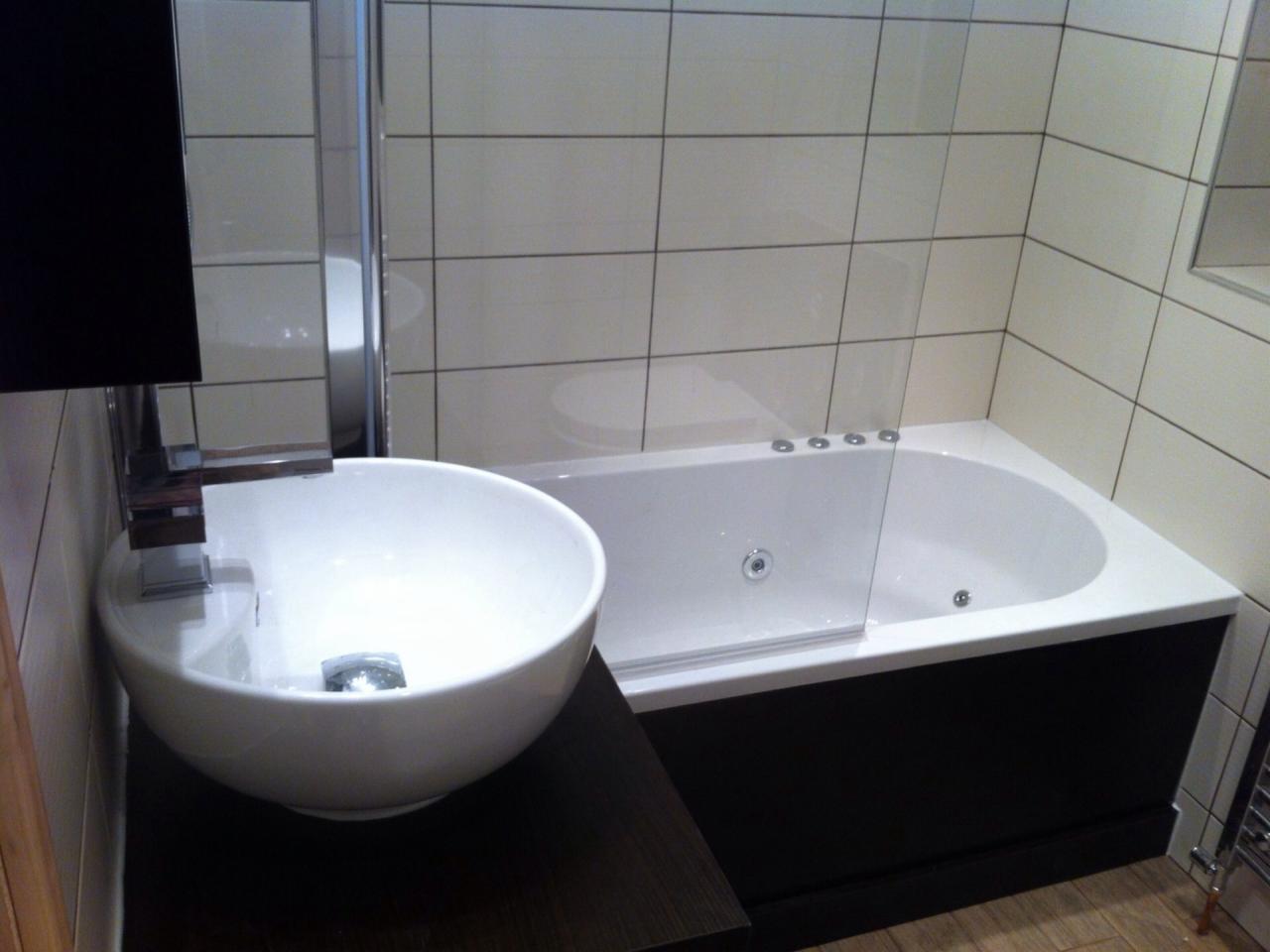 Nowoczesna łazienka z owalną umywalką na ciemnym blacie i białą wanną z hydromasażem, wykończona jasnymi płytkami.