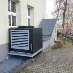 Zewnętrzna jednostka pompy ciepła Gorenje, zamontowana na metalowej platformie przy budynku z oknami, widoczna kratka wentylacyjna i logo firmy.