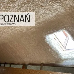 Poddasze pokryte grubą warstwą izolacji natryskowej z pianki, widoczne okno dachowe i logo firmy izolacyjnej.