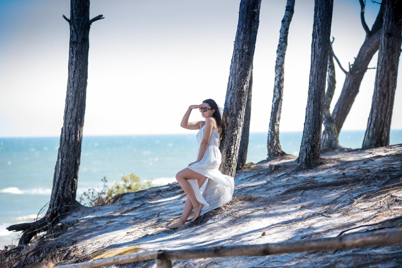 Kobieta w zwiewnej białej sukience, oparta o pień drzewa na klifie z widokiem na morze, w okularach przeciwsłonecznych, zasłaniająca dłonią oczy przed słońcem.