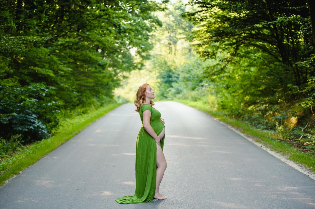 Ciężarna kobieta w zielonej sukni z rozcięciem, stojąca boso na asfalcie leśnej drogi, z zamkniętymi oczami i dłońmi na brzuchu.