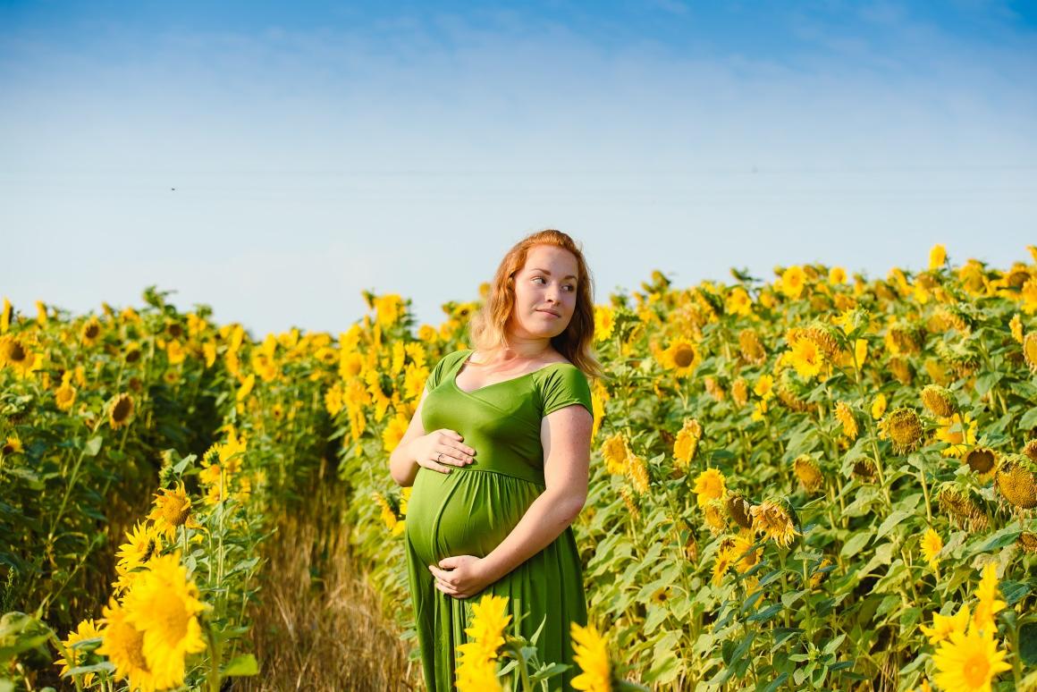 Kobieta w zielonej sukience ciążowej pozuje na tle pola słoneczników z delikatnym uśmiechem.