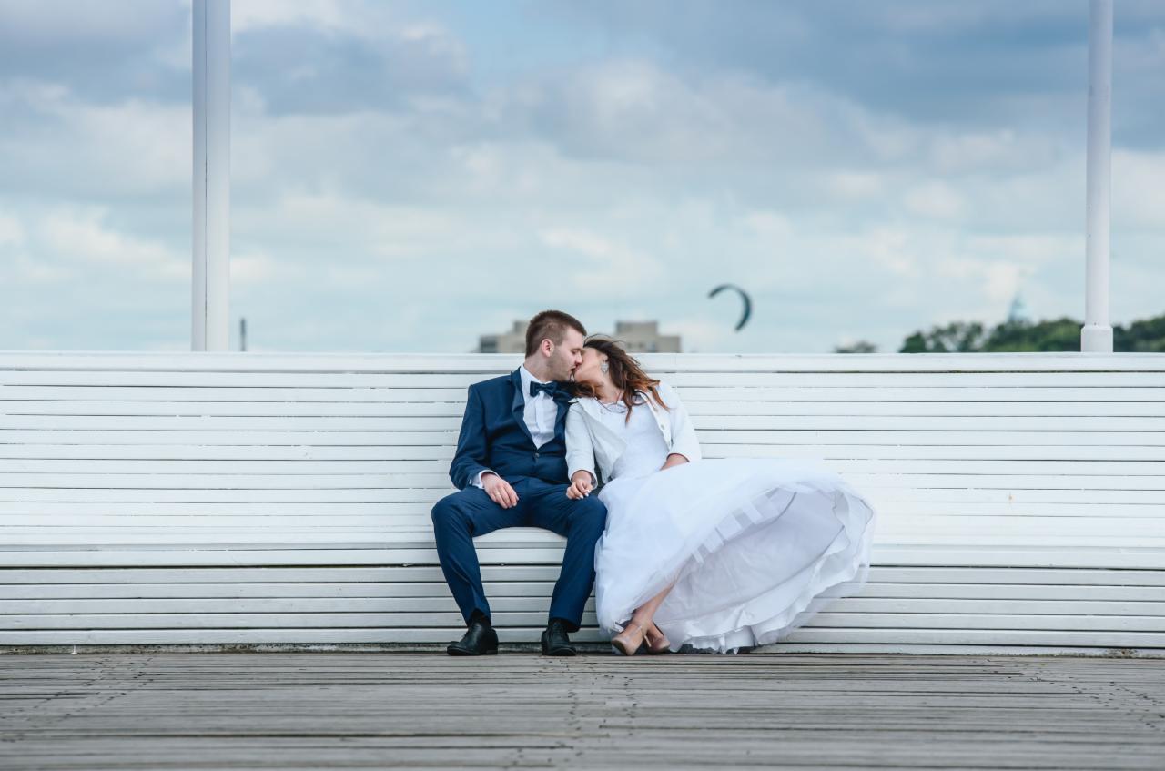 Para młoda w strojach ślubnych siedzi na białej ławce na molo, całując się na tle pochmurnego nieba i budynku w oddali.