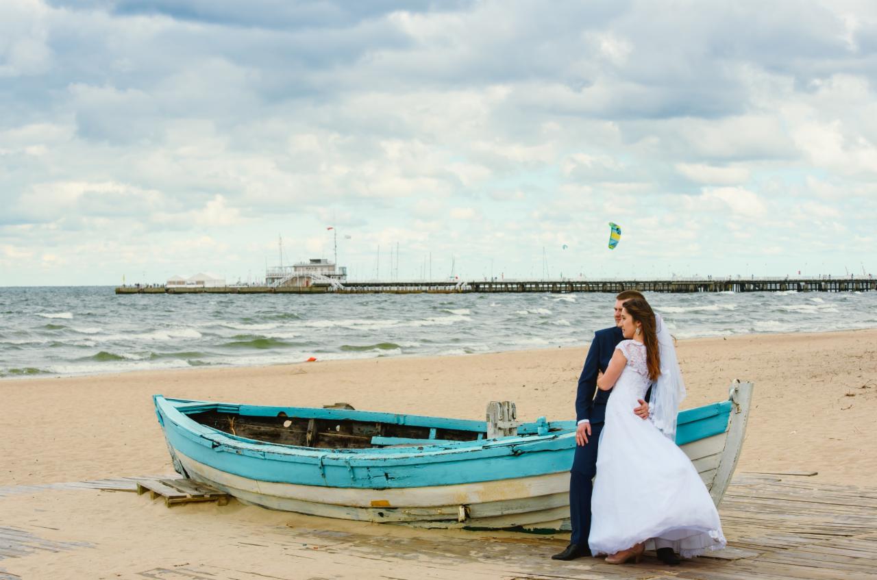 Para młoda w sukni ślubnej i garniturze pozuje na plaży obok starej, błękitnej łodzi, w tle molo i morze.
