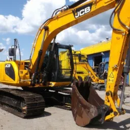 Żółta, gąsienicowa maszyna budowlana JCB z łyżką z przodu, zaparkowana na placu budowy, z innymi maszynami w tle i budynkiem z niebieskimi drzwiami.