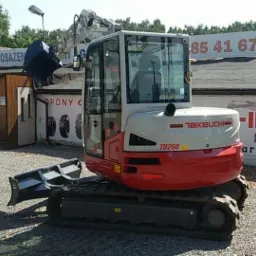 Kompaktowa, biało-czerwona maszyna Takeuchi TB260 na gąsienicach, zaparkowana przed budynkiem Komisu Wyposażenia, z łyżką skierowaną w lewo.
