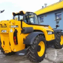 Żółta maszyna budowlana JCB 535-95 z podnośnikiem teleskopowym, widok z tyłu, na tle niebieskiego budynku z żółtym dachem.