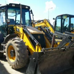 Żółta koparko-ładowarka JCB z widocznym osprzętem, zaparkowana obok innych maszyn budowlanych na placu składowym w słoneczny dzień.