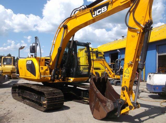 Żółta, gąsienicowa maszyna budowlana JCB z łyżką z przodu, zaparkowana na placu budowy, z innymi maszynami w tle i budynkiem z niebieskimi drzwiami.