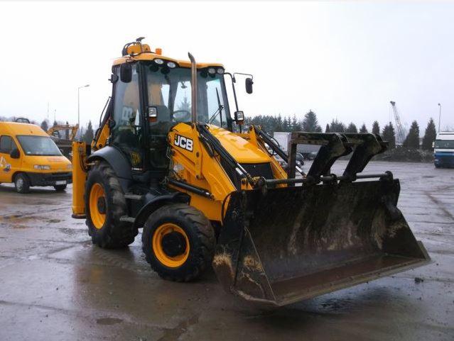 Żółta koparko-ładowarka JCB na placu budowy, widoczny przedni łyżka i kabina operatora, w tle żółty bus i dźwig.
