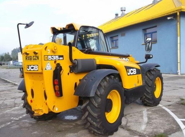Żółta maszyna budowlana JCB 535-95 z podnośnikiem teleskopowym, widok z tyłu, na tle niebieskiego budynku z żółtym dachem.