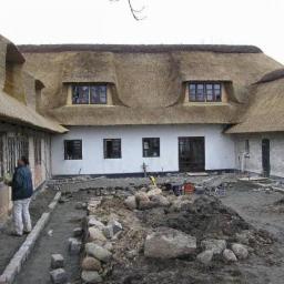 Cmj Design Totalentreprise - Tradycyjny dom z krytym strzechą dachem w trakcie prac wykończeniowych na zewnątrz, widoczny pracownik przy oknie i przygotowany teren pod ścieżkę.