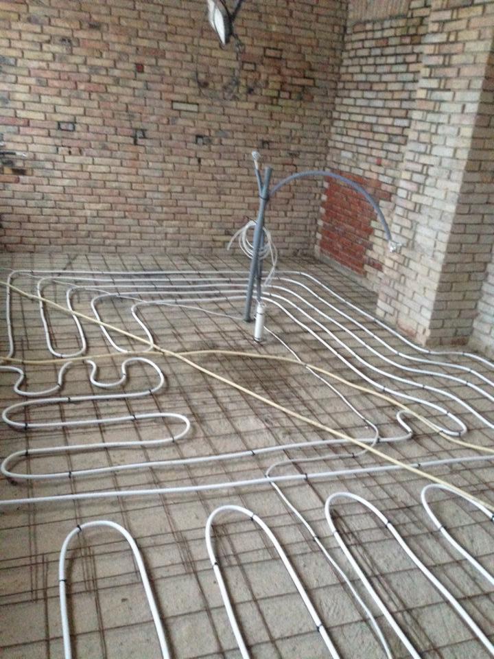 Instalacja ogrzewania podłogowego z rur w pomieszczeniu z surową cegłą, widoczne mocowania do siatki zbrojeniowej.