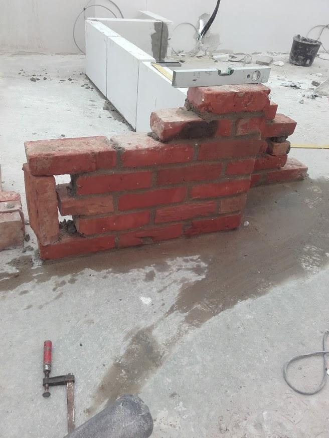 Wznoszenie fragmentu muru z czerwonej cegły na mokrej, betonowej posadzce w trakcie prac budowlanych, widoczny poziomica i biała konstrukcja w tle.