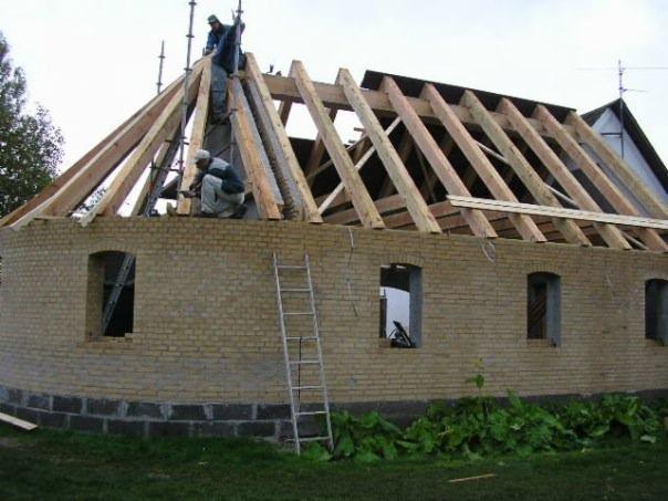 Budowa dachu na okrągłym budynku z żółtej cegły. Dwóch robotników pracuje na konstrukcji z drewnianych belek, widoczna drabina oparta o ścianę i niezabudowane otwory okienne.