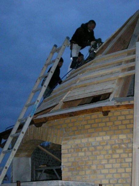 Ekipa budowlana pracuje na dachu rozbudowywanego domu z żółtej cegły, widoczna drabina i narzędzia.