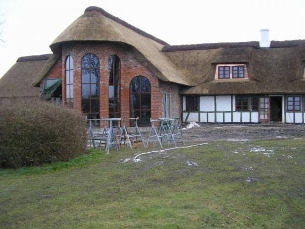 Dom z cegły z charakterystycznym strzechą na dachu, widoczne rusztowania i teren w trakcie prac wykończeniowych.