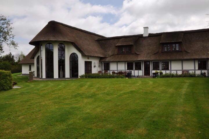 Biały dom z strzechą i dużymi, łukowatymi oknami, otoczony rozległym, równo przystrzyżonym trawnikiem.