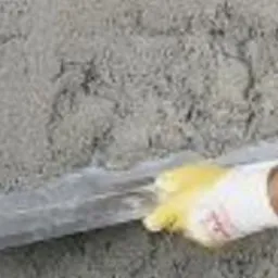 Nakładanie zaprawy na ścianę za pomocą długiej, metalowej pacy. Ręka w żółtej rękawicy roboczej. Widoczna surowa, nierówna powierzchnia ściany w szarym kolorze.