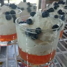 Deser w szklankach: pomarańczowa galaretka, biała pianka i borówki, ułożony na metalowej półce chłodziarki.