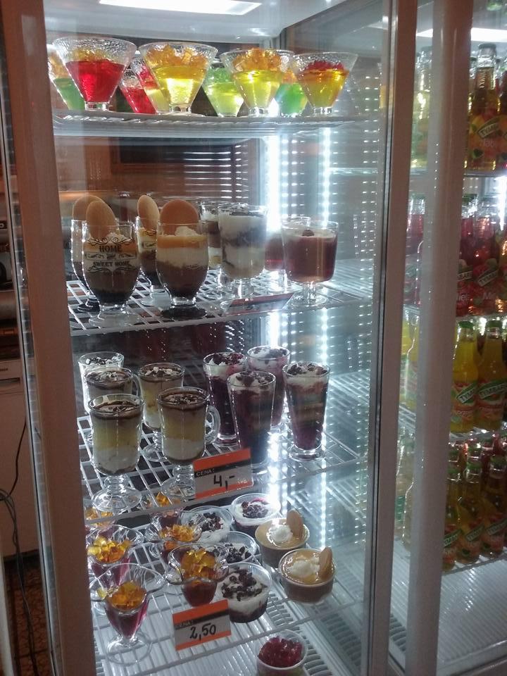 Szklana witryna chłodnicza wypełniona różnorodnymi deserami: galaretki w kieliszkach, kremy warstwowe, puddingi z owocami, ciasteczkami i posypką czekoladową, obok butelki napojów, widoczne...