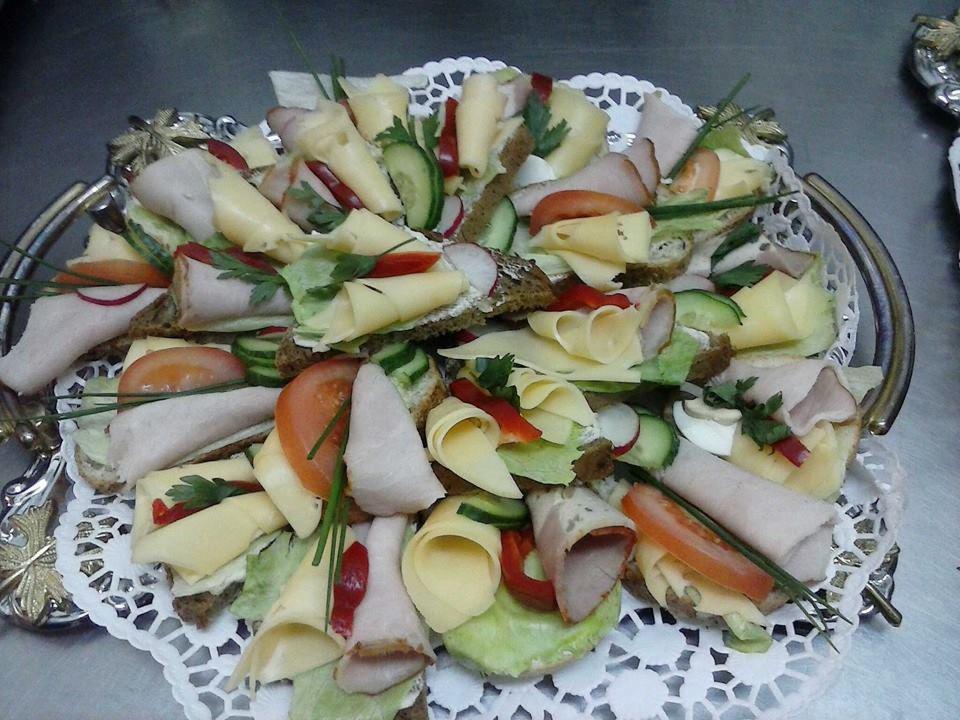 Dekoracyjna srebrna patera z różnorodnymi kanapkami: ciemne pieczywo, ser, wędlina, warzywa (ogórek, rzodkiewka, pomidor, papryka), sałata i zioła, ułożonymi na białej koronkowej serwetce.