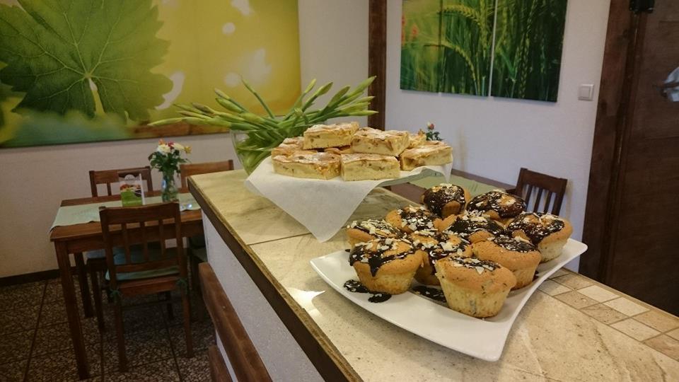 Blat z jasnego kamienia z ekspozycją ciast: pokrojona szarlotka na podwyższeniu i muffiny polane czekoladą na białym talerzu; w tle stół nakryty zielonym obrusem i kwiaty w wazonie, a na ścianie...