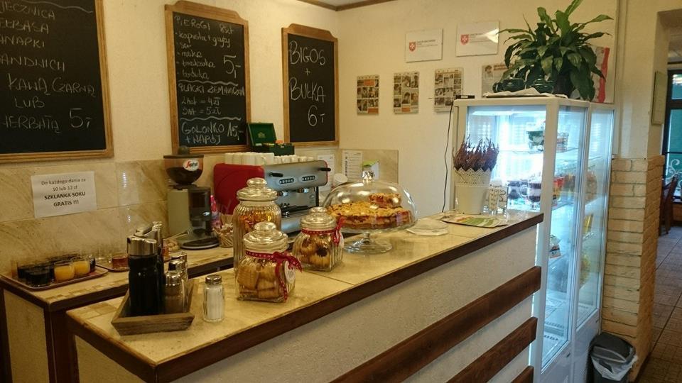 Wnętrze lokalu gastronomicznego: lada z ciastem pod kloszem, słoikami z ciasteczkami, ekspres do kawy, napoje oraz tablice z menu wypisanym kredą.