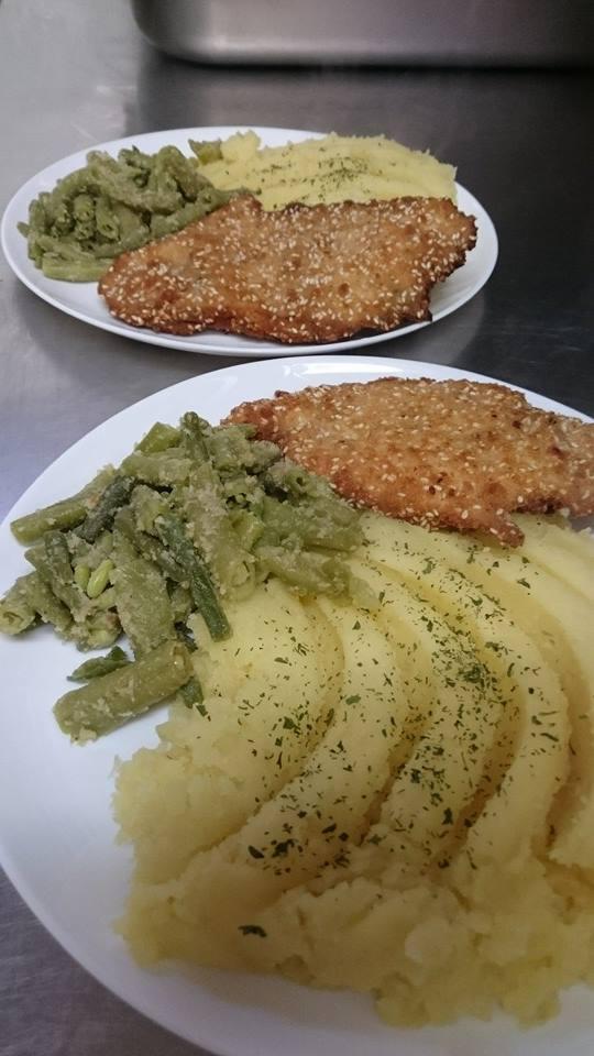 Dwa talerze z obiadem: kotlet w panierce sezamowej, puree ziemniaczane posypane ziołami i fasolka szparagowa z sosem, na metalowym blacie.