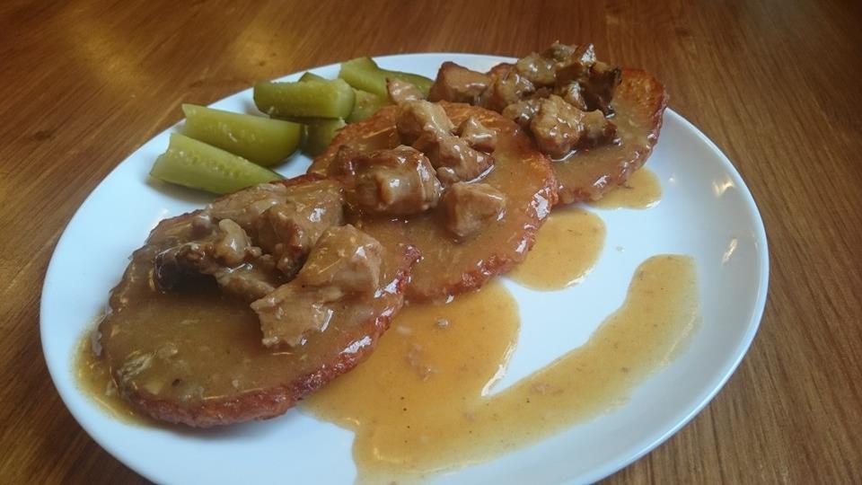 Trzy placki ziemniaczane polane sosem z kawałkami mięsa, podane z ogórkami kiszonymi na białym talerzu, ułożonym na drewnianym stole.