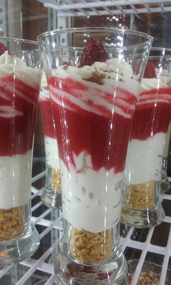 Deser warstwowy w szklance: spód z pokruszonych ciastek, kremowa warstwa, mus truskawkowy z kleksem bitej śmietany i świeżą truskawką na wierzchu, posypany brązowym proszkiem.