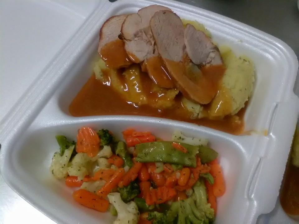 Porcja obiadowa w białym pojemniku styropianowym: plasterki pieczonego mięsa polane sosem na puree ziemniaczanym, obok mieszanka gotowanych warzyw, w tym marchewka i brokuły.