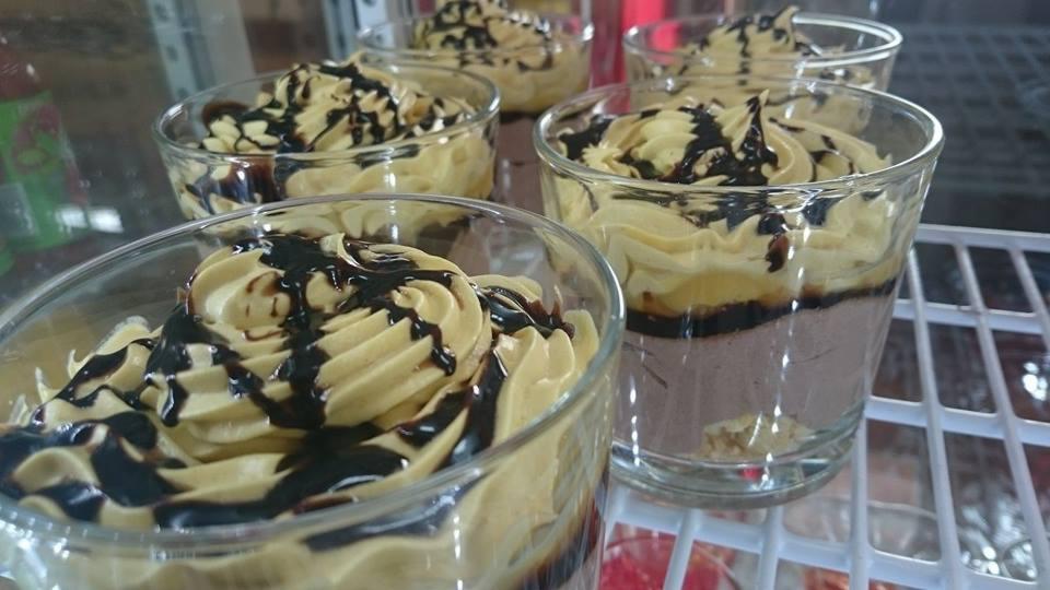 Deser w szklanych naczyniach z kremem, polewą czekoladową i widocznymi warstwami, ustawiony na białej półce chłodniczej.