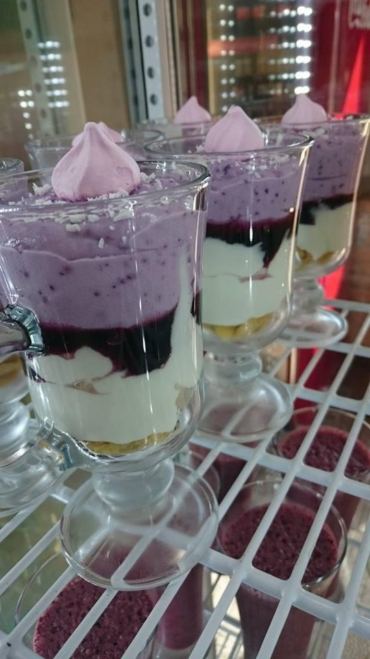 Szklanki z deserem warstwowym o fioletowych odcieniach, zwieńczone różową bezą i posypane wiórkami kokosowymi, ustawione na plastikowej kratce w chłodnym oświetleniu.