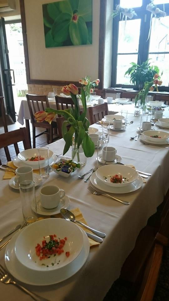 Długi stół nakryty białym obrusem z zastawą dla wielu osób, minimalistyczne dekoracje kwiatowe w wazonach, talerze z sałatką pomidorową, biała porcelana, drewniane krzesła, obraz z motywem...