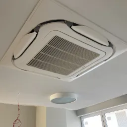 Kasetonowa jednostka klimatyzacji sufitowej w jasnym pomieszczeniu, widoczne wystające przewody elektryczne, okrągła lampa sufitowa w tle.