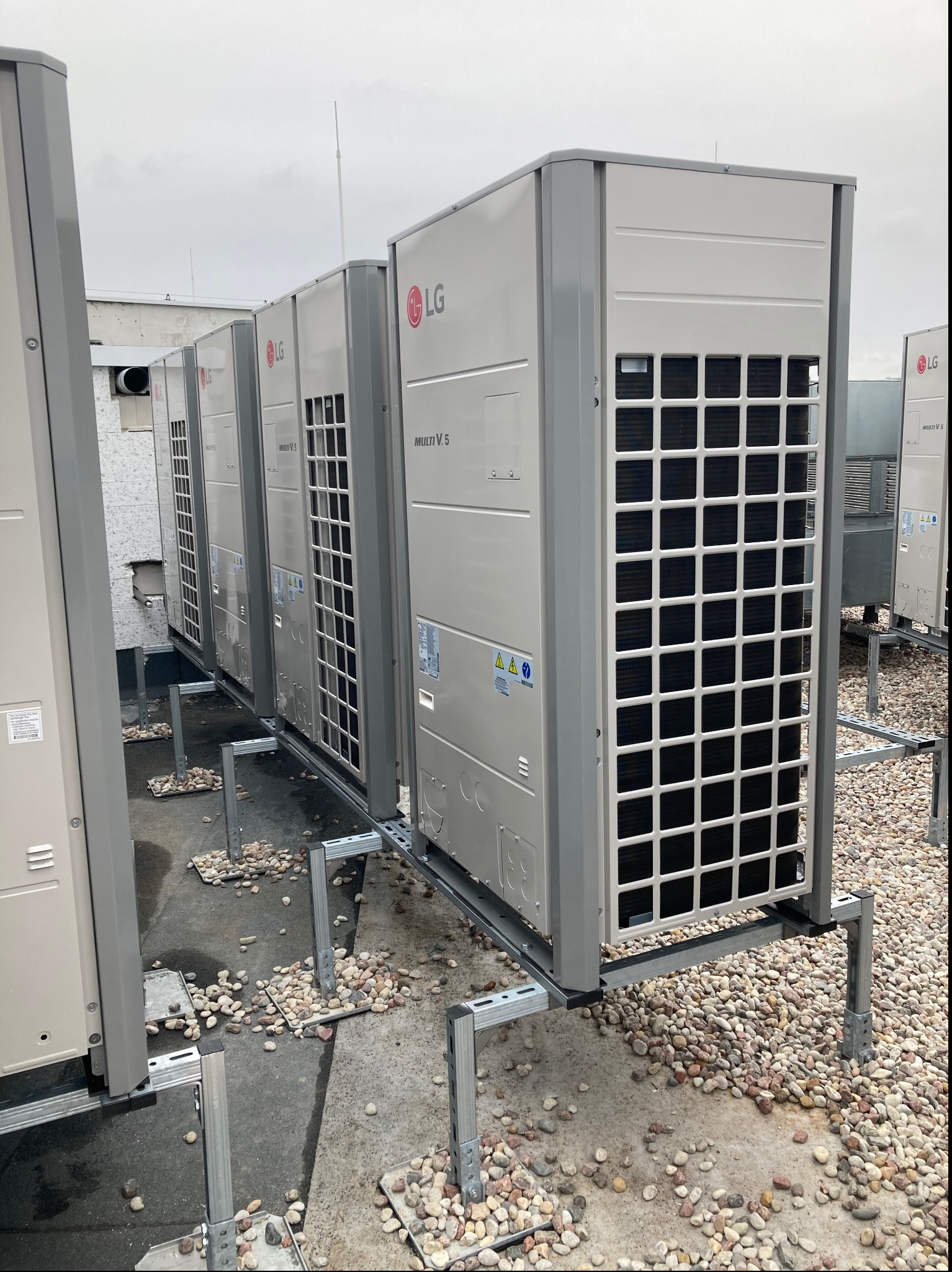 Rząd zewnętrznych jednostek klimatyzacyjnych LG Multi V 5 na dachu budynku komercyjnego, zamontowanych na metalowej konstrukcji z rozsypanymi kamieniami.
